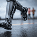 - VeyrZest.com Close-up of a wet robotic leg foot on the ground with blurred people in the background, suggesting a prosthetic or exoskeleton in use.
