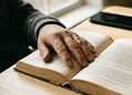 Close-up of a hand with a ring turning the pages of an open, old book on a wooden table, with a smartphone in the background.