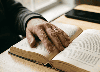 Close-up of a hand with a ring turning the pages of an open, old book on a wooden table, with a smartphone in the background.