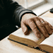 A_close-up_photograph_of_a_per_Nano_Banana_Pro_63984 (1) - VeyrZest.com Close-up of a hand with a ring turning the pages of an open, old book on a wooden table, with a smartphone in the background.