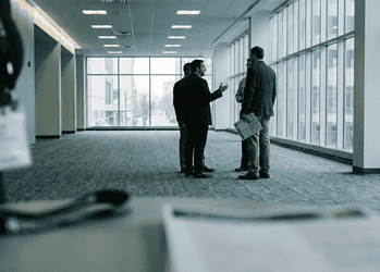 Group of business professionals conversing in a bright office hallway, one gesturing as they talk, with a blurred desk and badge in the foreground.