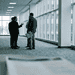 A_conference_corridor_or_hallw_Nano_Banana_Pro_75690 (1) - VeyrZest.com Group of business professionals conversing in a bright office hallway, one gesturing as they talk, with a blurred desk and badge in the foreground.
