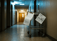 Dim hospital hallway with blue-tinted lighting and a metal cart holding papers and clipboards in the foreground