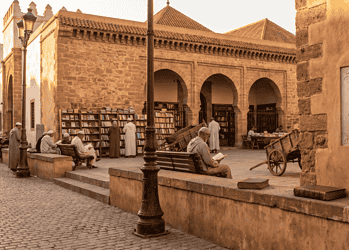 Old stone courtyard with arches, bookshelves, and men reading outdoors at a historic library market during sunset.