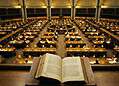 Open book on a lectern overlooking a grand library reading room filled with desks and warm lamps.