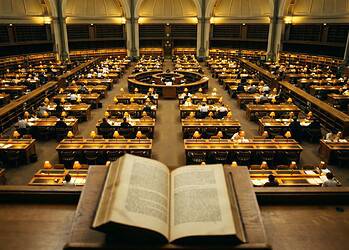 Open book on a lectern overlooking a grand library reading room filled with desks and warm lamps.