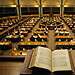 Open book on a lectern overlooking a grand library reading room filled with desks and warm lamps.