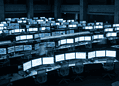 Trading floor with rows of monitors showing stock charts and graphs; a large clock on the wall.