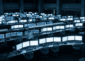 Trading floor with rows of monitors showing stock charts and graphs; a large clock on the wall.