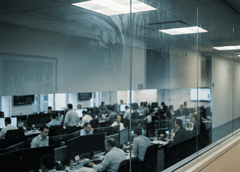 Open-plan office seen through a glass partition, with employees at desks and computer monitors.