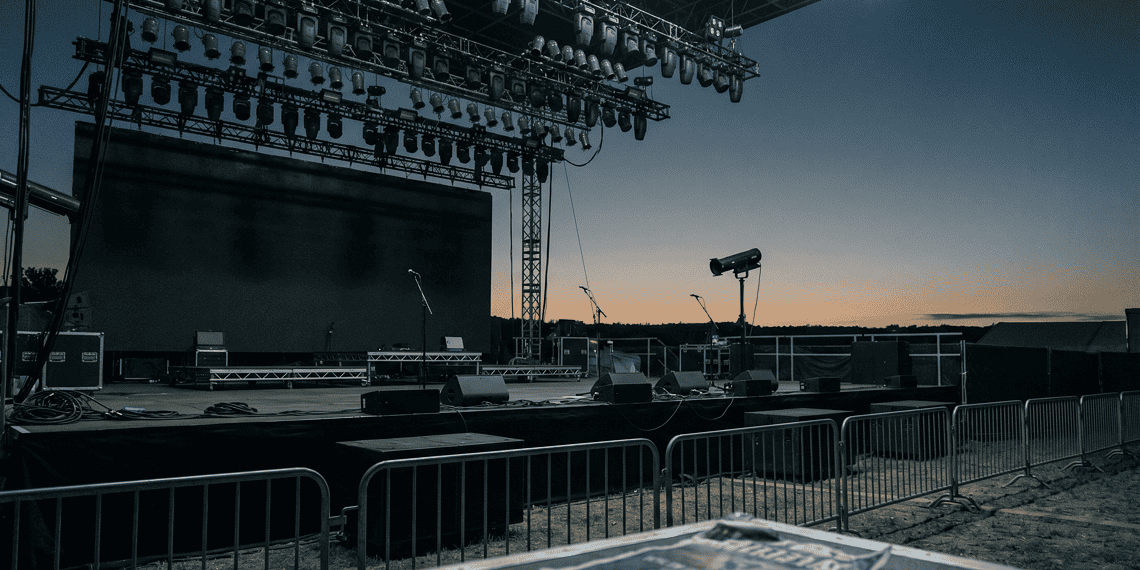 Outdoor concert stage at dusk with rigging and stage lights overhead, empty microphones on stands, and barricades in the foreground.