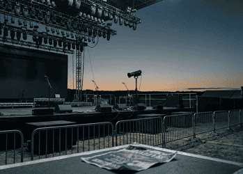 Outdoor concert stage at dusk with rigging and stage lights overhead, empty microphones on stands, and barricades in the foreground.