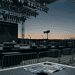 Outdoor concert stage at dusk with rigging and stage lights overhead, empty microphones on stands, and barricades in the foreground.