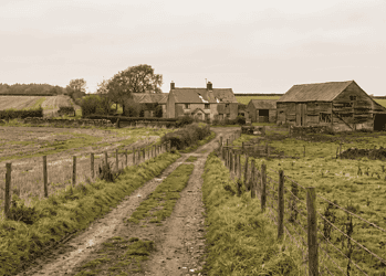 Dirt country road leads to a stone farmhouse with a weathered barn on a grassy farm under an overcast sky.