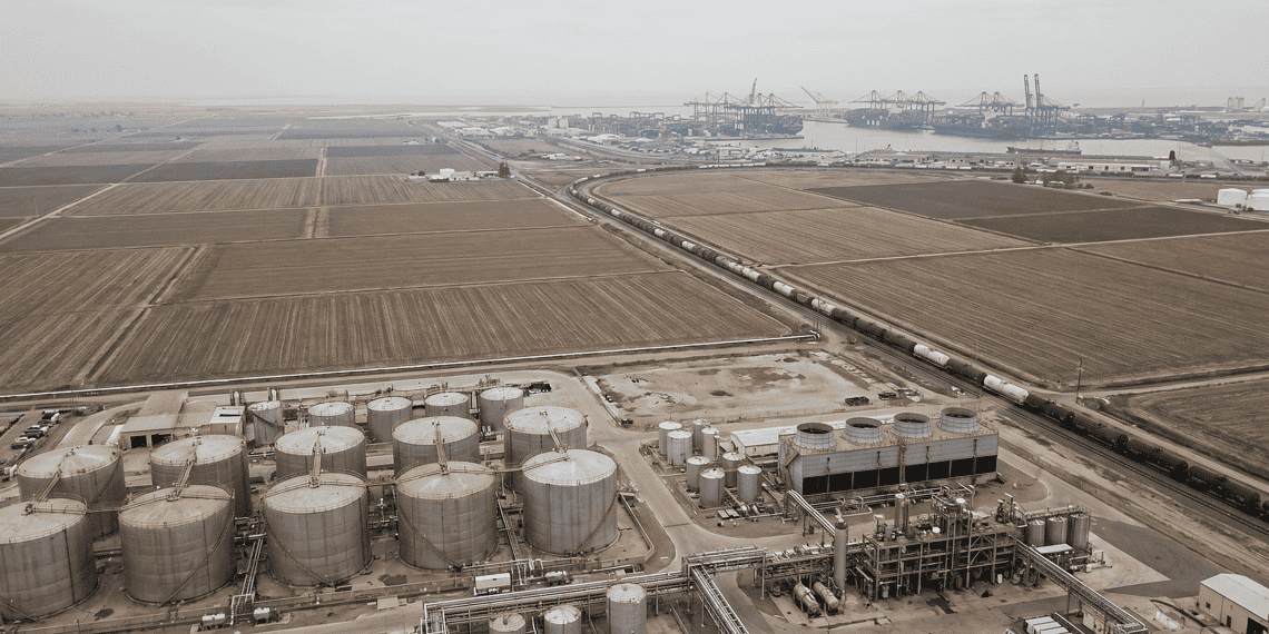 - VeyrZest.com Aerial view of an oil refinery with large cylindrical storage tanks in the foreground and a distant harbor with cranes on the horizon.