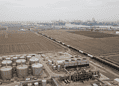 Aerial view of an oil refinery with large cylindrical storage tanks in the foreground and a distant harbor with cranes on the horizon.
