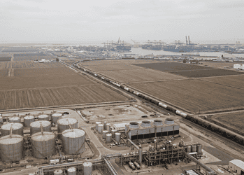 Aerial view of an oil refinery with large cylindrical storage tanks in the foreground and a distant harbor with cranes on the horizon.