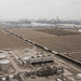- VeyrZest.com Aerial view of an oil refinery with large cylindrical storage tanks in the foreground and a distant harbor with cranes on the horizon.