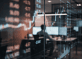 Trading room with glass walls and multiple screens showing stock charts and numbers, a blurred silhouette at a desk in the background.