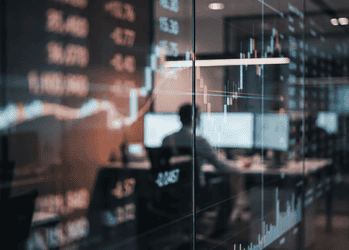 Trading room with glass walls and multiple screens showing stock charts and numbers, a blurred silhouette at a desk in the background.