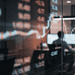 - VeyrZest.com Trading room with glass walls and multiple screens showing stock charts and numbers, a blurred silhouette at a desk in the background.