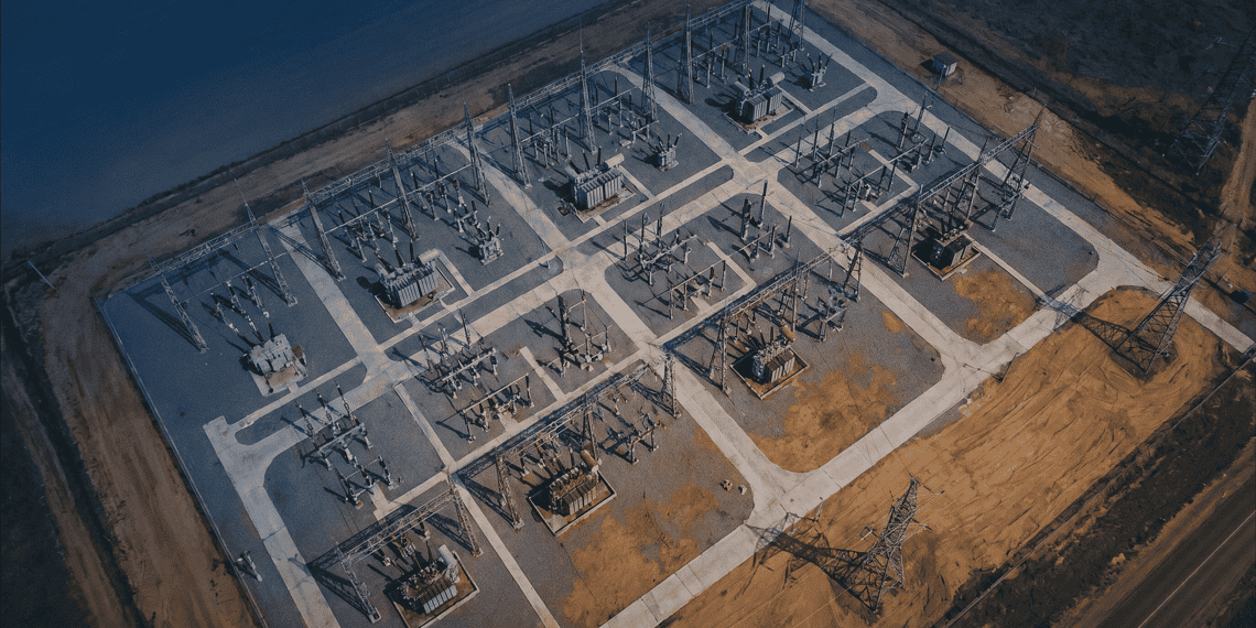 - VeyrZest.com Aerial view of an electrical substation with transformers and high‑voltage lines arranged in a geometric grid on a paved site beside a barren landscape.