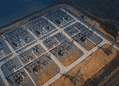 Aerial view of an electrical substation with transformers and high‑voltage lines arranged in a geometric grid on a paved site beside a barren landscape.
