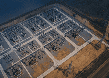 - VeyrZest.com Aerial view of an electrical substation with transformers and high‑voltage lines arranged in a geometric grid on a paved site beside a barren landscape.