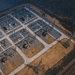 - VeyrZest.com Aerial view of an electrical substation with transformers and high‑voltage lines arranged in a geometric grid on a paved site beside a barren landscape.