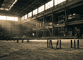 Industrial warehouse interior with scaffolding and metal framework, sepia-toned light streaming through high windows; foreground shows vertical pipes sticking from the dusty floor.