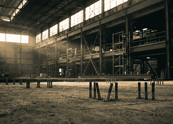 Industrial warehouse interior with scaffolding and metal framework, sepia-toned light streaming through high windows; foreground shows vertical pipes sticking from the dusty floor.