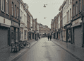 Quiet, wet street in a European town with closed shopfronts, benches and bicycles on the left, two people walking in the distance.