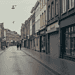 IMAGE_PROMPT_Wide-angle_photog_Nano_Banana_79562 - VeyrZest.com Quiet, wet street in a European town with closed shopfronts, benches and bicycles on the left, two people walking in the distance.