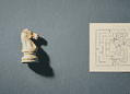 Beige chess knight model on the left casting a shadow, next to a beige notebook page with a black hand-drawn maze against a blue textured background.