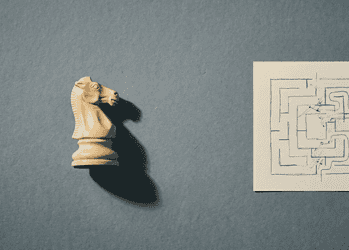 Beige chess knight model on the left casting a shadow, next to a beige notebook page with a black hand-drawn maze against a blue textured background.