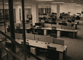 Open-plan office with rows of cubicles and desks, several chairs, and glass barriers in the foreground; no people present.