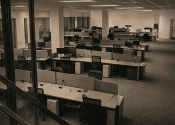 Open-plan office with rows of cubicles and desks, several chairs, and glass barriers in the foreground; no people present.