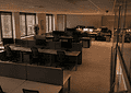 Open-plan office with rows of desks, computer monitors, and cubicle partitions under warm overhead lighting.