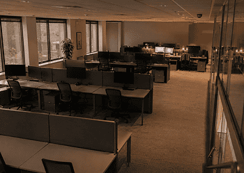 Open-plan office with rows of desks, computer monitors, and cubicle partitions under warm overhead lighting.