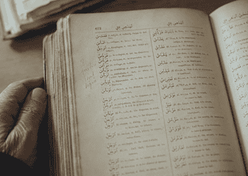 Open antique dictionary with Arabic entries, held by a left hand.