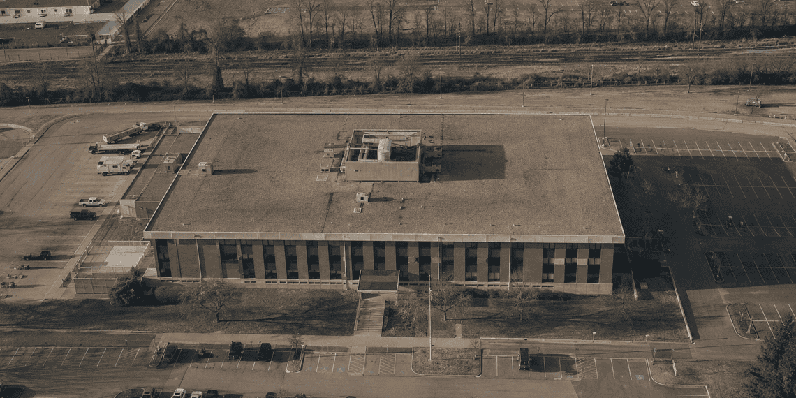 - VeyrZest.com Aerial view of a large rectangular office building with a flat roof, surrounded by parking lots and roads steamrollering around it.