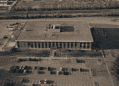 Aerial view of a large rectangular office building with a flat roof, surrounded by parking lots and roads steamrollering around it.