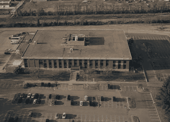 Aerial view of a large rectangular office building with a flat roof, surrounded by parking lots and roads steamrollering around it.