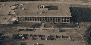 Aerial view of a large rectangular office building with a flat roof, surrounded by parking lots and roads steamrollering around it.